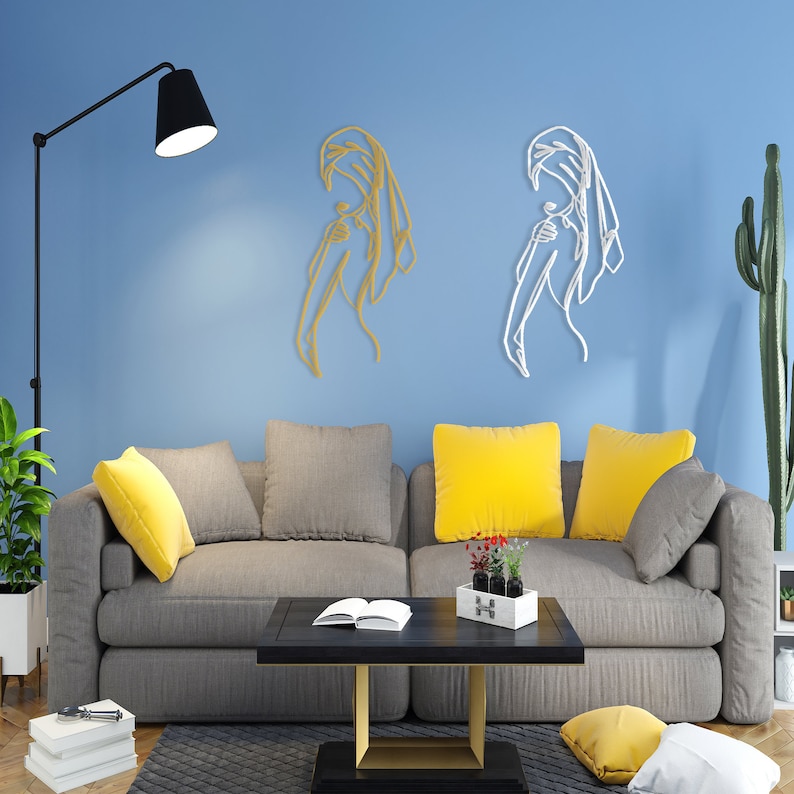 May include: Two metal wall sculptures of a woman in a flowing robe, one in gold and one in white, against a light blue wall. The sculptures are displayed above a gray sofa with yellow throw pillows.