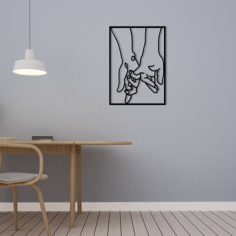 May include: Black metal wall art depicting two hands holding each other. The art is framed in a square black frame and is hanging on a light gray wall.