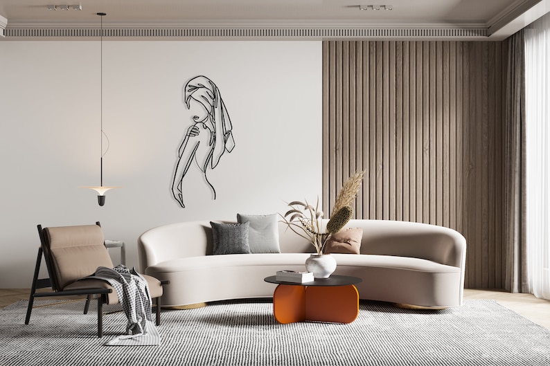 May include: A white curved sofa with a black metal coffee table and a black and white patterned rug. The coffee table has a unique orange base. A black line art drawing of a person with a towel on their head is on the wall behind the sofa.