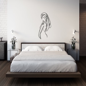 May include: A black metal wall art sculpture depicting a woman in a single line drawing. The sculpture is hanging on a white wall in a bedroom setting with a bed, nightstands, and a couch.