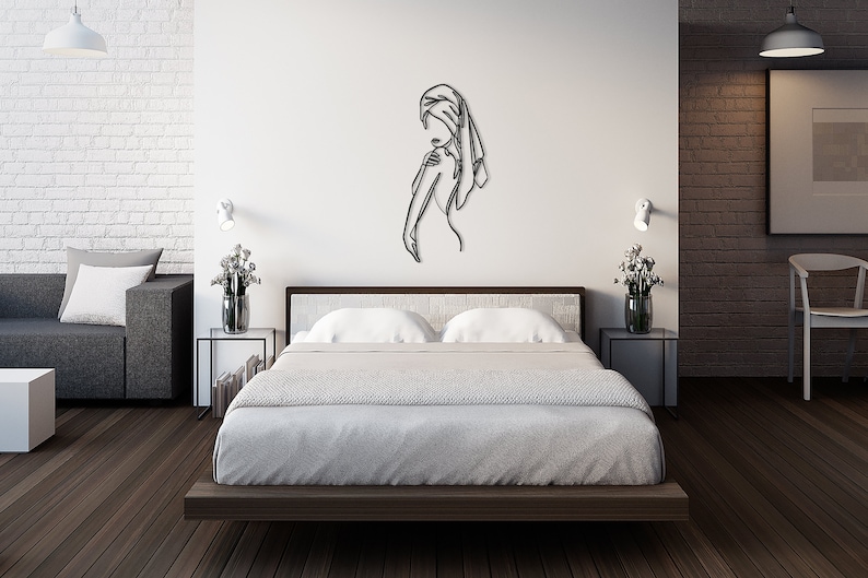 May include: A black metal wall art sculpture depicting a woman in a single line drawing. The sculpture is hanging on a white wall in a bedroom setting with a bed, nightstands, and a couch.
