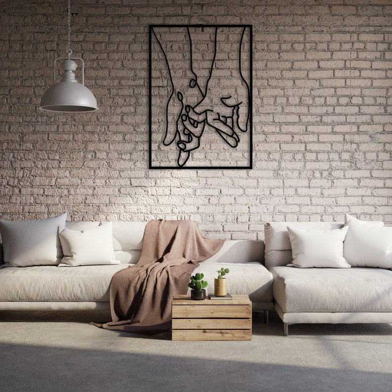 May include: A black metal wall art sculpture depicting two hands holding each other. The sculpture is mounted on a white brick wall above a white couch with brown throw blankets.