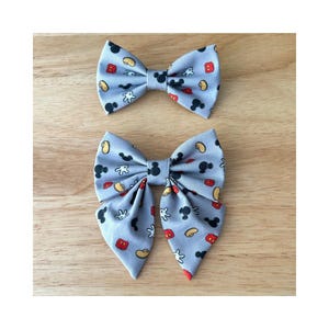 Peut inclure: Deux nœuds papillon gris avec un imprimé Mickey Mouse. Le plus petit nœud papillon a des icônes Mickey Mouse rouges, jaunes et blanches. Le plus grand nœud papillon a un imprimé plus détaillé avec des icônes Mickey Mouse rouges, jaunes, blanches et noires.