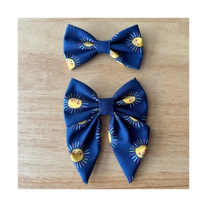 May include: Two navy blue hair bows with a yellow sun pattern. The smaller bow is tied in a traditional bow shape, while the larger bow is tied in a more relaxed, oversized style.