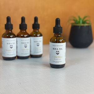 May include: Four brown glass bottles with droppers, labeled "Rub & Tug" and "Come Hither Beard Oil". The bottles are on a white surface with a plant in a pot in the background.