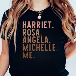 Könnte beinhalten: Schwarzes T-Shirt mit den Namen "Harriet, Rosa, Angela, Michelle, Me" in kupferfarbener Schrift. Das Shirt hat einen Rundhalsausschnitt und kurze Ärmel.