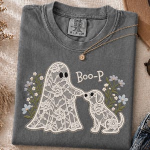 Op de afbeelding: Een antracietgrijs t-shirt met ronde hals voorzien van een witte geest en hond met kantpatroon en de tekst Boo-P. Het ontwerp bevat blauwe en groene bloemaccenten. Het t-shirt is voorzien van een Comfort Colors label.