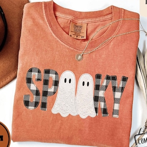 May include: A burnt orange short-sleeve t-shirt featuring the word SPOOKY in black and white buffalo plaid lettering, with two white ghost figures replacing the letter O. The shirt has a crew neckline and a Comfort Colors brand label.