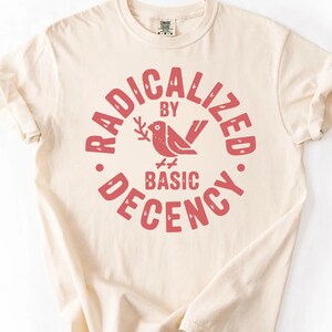 Puede incluir: Camiseta color crema con mangas enrolladas. La parte delantera presenta un diseño circular rojo con las palabras "RADICALIZED BY BASIC DECENCY" alrededor de un pájaro que sostiene una rama.