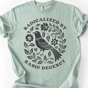 Può includere: Una maglietta verde chiaro con la scritta "RADICALIZED BY BASIC DECENCY" in un disegno circolare. Il disegno presenta un uccello nero circondato da fiori e foglie. La maglietta ha le maniche arrotolate.
