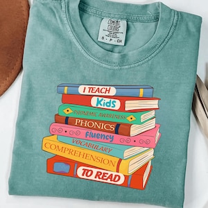 May include: A teal Comfort Colors t-shirt featuring a stack of colorful books. Each book has a title related to reading, such as "I Teach Kids," "Phonics," and "Vocabulary." The shirt is a comfortable, casual style.