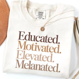 Könnte beinhalten: Cremefarbenes T-Shirt mit den Worten "Educated. Motivated. Elevated. Melanated." in braunen und beigen Buchstaben. Eine goldene Halskette mit Anhänger liegt auf dem Shirt. Ein brauner Hut und getrocknete Palmblätter sind im Hintergrund.