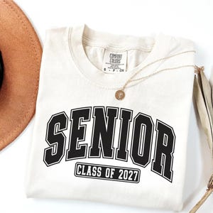 Puede incluir: Una camiseta de manga corta de color crema con la palabra SENIOR en letras negras grandes y el texto CLASS OF 2027 dentro de un rect&aacute;ngulo negro debajo. La camiseta tiene cuello redondo y una etiqueta de la marca Comfort Colors.