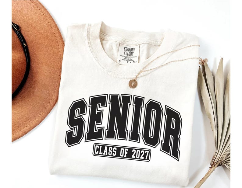 Puede incluir: Una camiseta de manga corta de color crema con la palabra SENIOR en letras negras grandes y el texto CLASS OF 2027 dentro de un rect&aacute;ngulo negro debajo. La camiseta tiene cuello redondo y una etiqueta de la marca Comfort Colors.