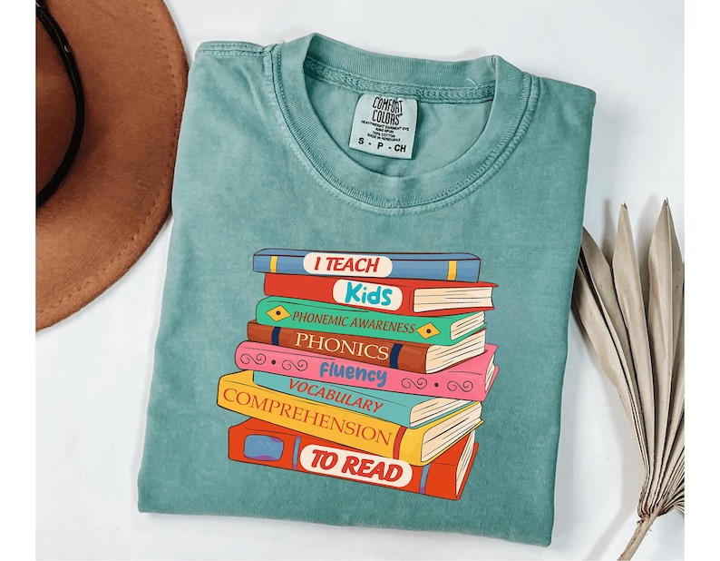 May include: A teal Comfort Colors t-shirt featuring a stack of colorful books. Each book has a title related to reading, such as "I Teach Kids," "Phonics," and "Vocabulary." The shirt is a comfortable, casual style.