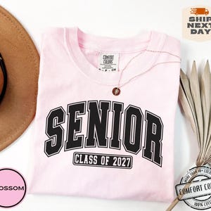Puede incluir: Una camiseta de manga corta de color rosa claro con la palabra SENIOR en letras negras grandes con borde blanco, y el texto CLASS OF 2027 en un marco rectangular negro debajo. La camiseta tiene una etiqueta de la marca Comfort Colors.
