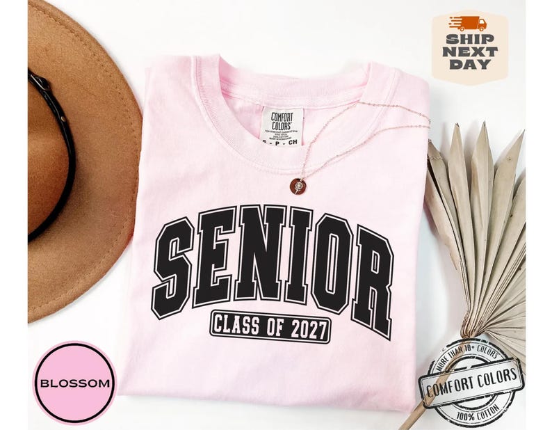 Puede incluir: Una camiseta de manga corta de color rosa claro con la palabra SENIOR en letras negras grandes con borde blanco, y el texto CLASS OF 2027 en un marco rectangular negro debajo. La camiseta tiene una etiqueta de la marca Comfort Colors.