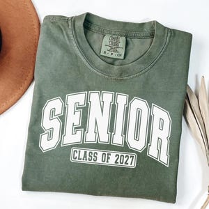 Puede incluir: Una camiseta de manga corta color verde oliva con cuello redondo. El frente presenta la palabra SENIOR en letras may&uacute;sculas blancas grandes con un borde blanco, y debajo, el texto CLASS OF 2027 dentro de un borde rectangular blanco.