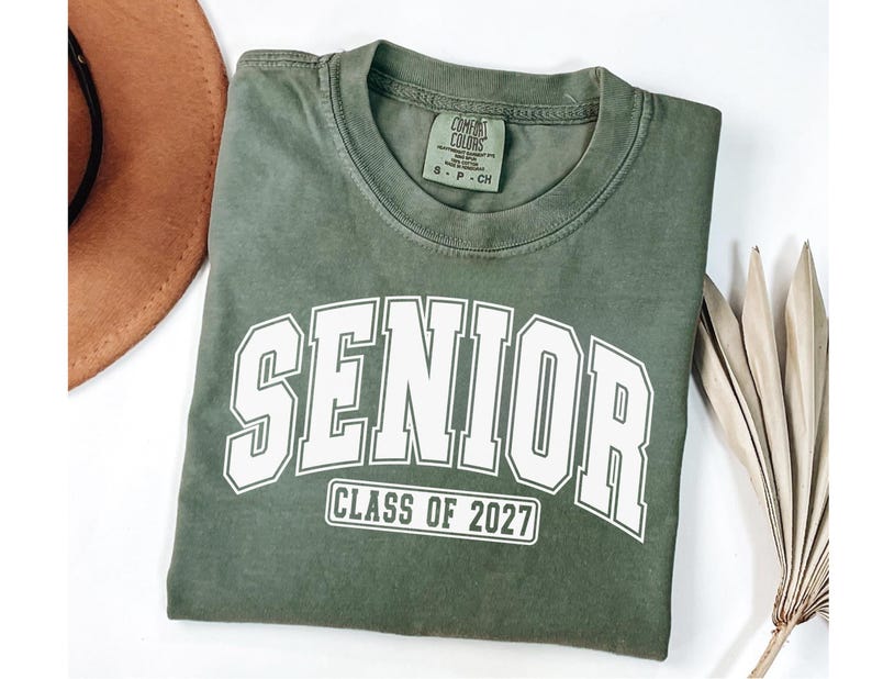Puede incluir: Una camiseta de manga corta color verde oliva con cuello redondo. El frente presenta la palabra SENIOR en letras may&uacute;sculas blancas grandes con un borde blanco, y debajo, el texto CLASS OF 2027 dentro de un borde rectangular blanco.