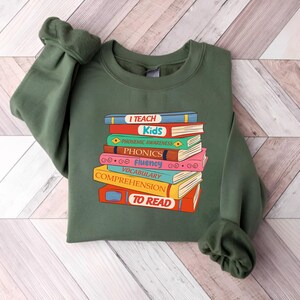 May include: An olive green sweatshirt featuring a stack of colorful books with the words "I Teach Kids" and other reading-related terms. The books are in various colors, including red, blue, and yellow. The sweatshirt has long sleeves.