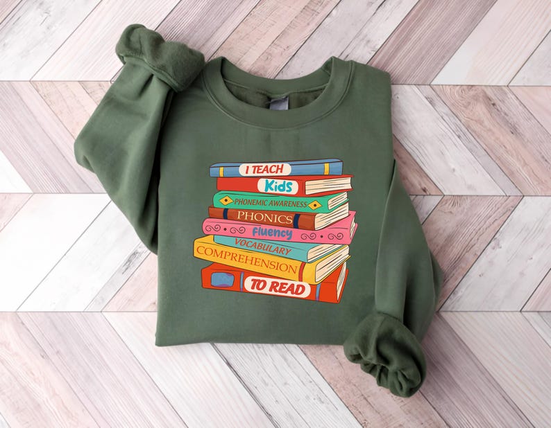 May include: An olive green sweatshirt featuring a stack of colorful books with the words "I Teach Kids" and other reading-related terms. The books are in various colors, including red, blue, and yellow. The sweatshirt has long sleeves.
