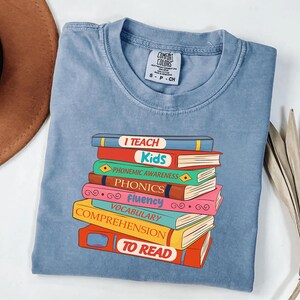 May include: A light blue Comfort Colors t-shirt featuring a stack of colorful books with the words "I TEACH Kids" and other reading-related terms. The shirt has a classic crew neck and short sleeves.
