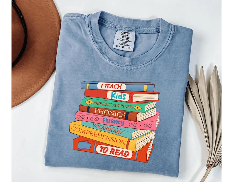 May include: A light blue Comfort Colors t-shirt featuring a stack of colorful books with the words "I TEACH Kids" and other reading-related terms. The shirt has a classic crew neck and short sleeves.