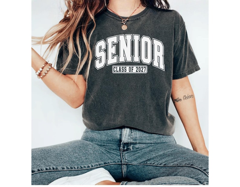 Puede incluir: Una camiseta de manga corta gris carb&oacute;n con el texto blanco SENIOR en una fuente universitaria y el texto CLASS OF 2027 debajo. La camiseta tiene un cuello redondo y un aspecto desgastado de inspiraci&oacute;n vintage combinada con pantalones vaqueros azules.