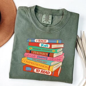 May include: Olive green t-shirt with a graphic of a stack of colorful books. Each book has a title like "I Teach Kids", "Phonemic Awareness", and "To Read". The shirt is made of a soft material and has a Comfort Colors tag.