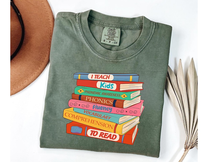 May include: Olive green t-shirt with a graphic of a stack of colorful books. Each book has a title like "I Teach Kids", "Phonemic Awareness", and "To Read". The shirt is made of a soft material and has a Comfort Colors tag.