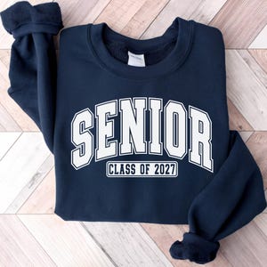 Puede incluir: Una sudadera azul marino de cuello redondo con la palabra SENIOR en una fuente blanca grande de estilo universitario arqueada en el pecho, con el texto CLASS OF 2027 impreso en un recuadro blanco m&aacute;s peque&ntilde;o justo debajo de las letras.