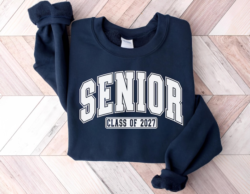 Puede incluir: Una sudadera azul marino de cuello redondo con la palabra SENIOR en una fuente blanca grande de estilo universitario arqueada en el pecho, con el texto CLASS OF 2027 impreso en un recuadro blanco m&aacute;s peque&ntilde;o justo debajo de las letras.