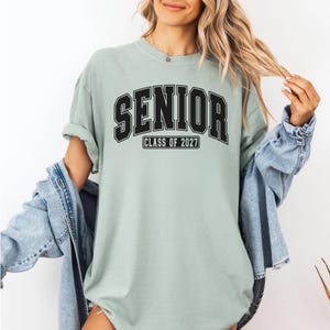 Puede incluir: Una camiseta de manga corta color verde salvia que presenta la palabra SENIOR en una fuente negra grande de estilo universitario, con el texto CLASS OF 2027 impreso en un recuadro rectangular negro m&aacute;s peque&ntilde;o justo debajo de las letras principales.