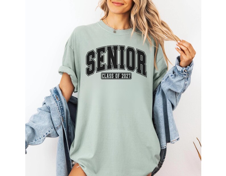 Puede incluir: Una camiseta de manga corta color verde salvia que presenta la palabra SENIOR en una fuente negra grande de estilo universitario, con el texto CLASS OF 2027 impreso en un recuadro rectangular negro m&aacute;s peque&ntilde;o justo debajo de las letras principales.