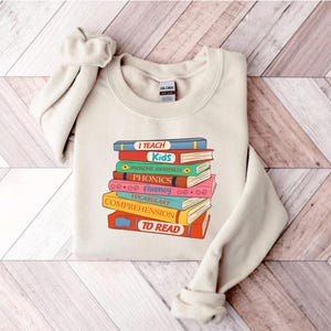 May include: A cream-colored sweatshirt featuring a stack of colorful books with the words "I TEACH Kids" and other reading-related terms. The design is in a retro style, with a vintage aesthetic. The sweatshirt is made of a soft material.