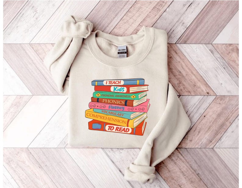 May include: A cream-colored sweatshirt featuring a stack of colorful books with the words "I TEACH Kids" and other reading-related terms. The design is in a retro style, with a vintage aesthetic. The sweatshirt is made of a soft material.