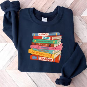 May include: Navy blue sweatshirt featuring a stack of colorful books with the words "I Teach Kids" and other reading-related terms. The books are in various colors, including red, blue, green, and pink. The sweatshirt has a crew neck and long sleeves.