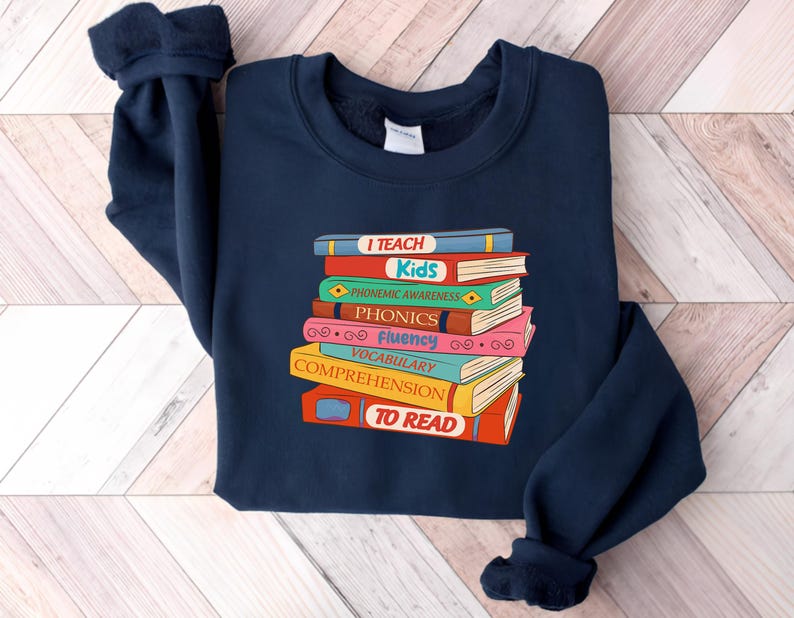 May include: Navy blue sweatshirt featuring a stack of colorful books with the words "I Teach Kids" and other reading-related terms. The books are in various colors, including red, blue, green, and pink. The sweatshirt has a crew neck and long sleeves.