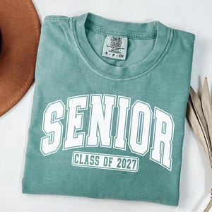Puede incluir: Una camiseta de manga corta color verde salvia con cuello redondo. El frente presenta letras blancas de estilo universitario que dicen SENIOR y un recuadro rectangular debajo con el texto CLASS OF 2027. Etiqueta interior visible.