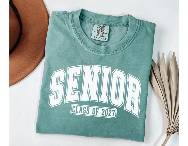 Puede incluir: Una camiseta de manga corta color verde salvia con cuello redondo. El frente presenta letras blancas de estilo universitario que dicen SENIOR y un recuadro rectangular debajo con el texto CLASS OF 2027. Etiqueta interior visible.