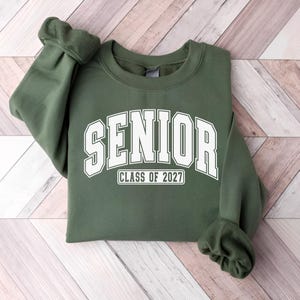 Puede incluir: Una sudadera de cuello redondo color verde oliva que presenta la palabra SENIOR en una fuente blanca grande de estilo universitario arqueada en el pecho, con el texto CLASS OF 2027 impreso en un recuadro blanco m&aacute;s peque&ntilde;o debajo.