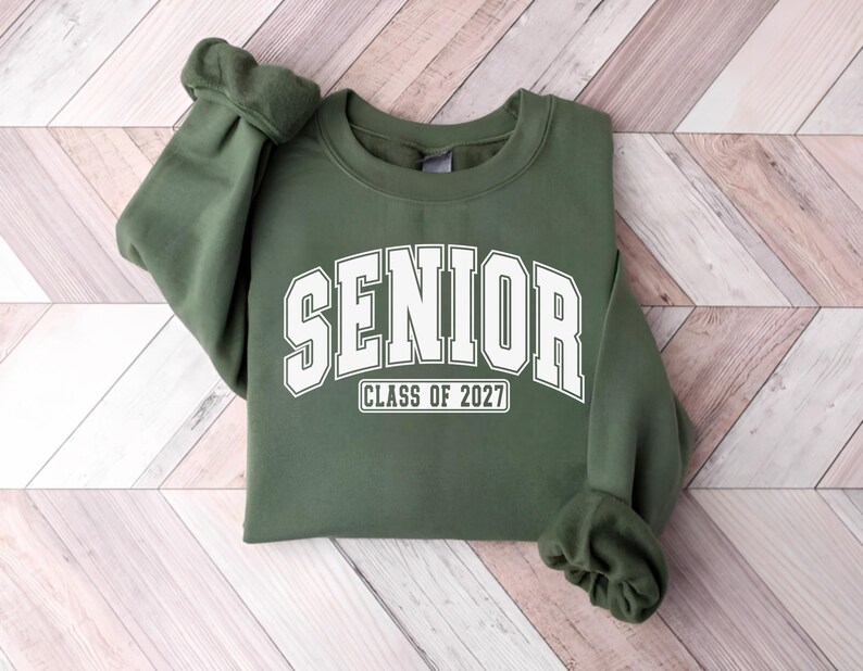 Puede incluir: Una sudadera de cuello redondo color verde oliva que presenta la palabra SENIOR en una fuente blanca grande de estilo universitario arqueada en el pecho, con el texto CLASS OF 2027 impreso en un recuadro blanco m&aacute;s peque&ntilde;o debajo.