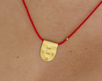 May include: A gold-toned pendant with a rounded bottom edge and a textured surface featuring a central cross-like motif surrounded by four small dots. The pendant is suspended from a thin, vibrant red cord necklace that includes a small gold bead.