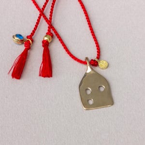 Könnte beinhalten: Eine goldfarbene Halskette mit Anhänger in einer einzigartigen Form, mit drei kreisförmigen Aussparungen. Die Halskette hat eine rote Kordel mit roten Quasten und einem kleinen blauen und goldenen Anhänger. Eine kleine goldene Scheibe ist ebenfalls an der Kordel befestigt.