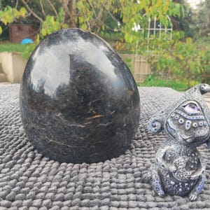 May include: A large, dark, egg-shaped stone with a polished surface sits next to a small, gray figurine with a whimsical design. The figurine has a large eye and decorative patterns. The objects are on a textured gray surface.