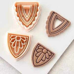 Puede incluir: Cuatro cortadores de galletas en forma de escudo. Dos son de plástico rosa claro y dos marrones. Un cortador rosa tiene un borde festoneado y un diseño anidado. El otro cortador rosa tiene un diseño floral. Los cortadores marrones tienen las mismas formas.