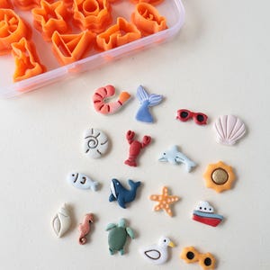 Puede incluir: Una colección de pequeños dijes de arcilla coloridos con formas temáticas del océano, que incluyen un camarón, cola de sirena, gafas de sol, conchas, langosta, delfín, ballena, estrella de mar, sol, barco, caballito de mar, tortuga y gaviota, junto con cortadores de galletas de plástico naranja.