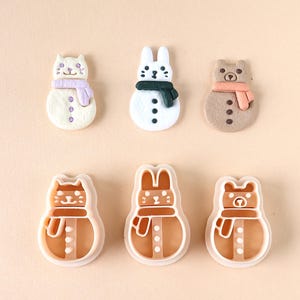 Op de afbeelding: Een verzameling van zes sneeuwmanvormige koekjessnijders en koekjes. De koekjes zijn versierd om op een kat, konijn en beer te lijken, elk met een sjaal. De snijders zijn gemaakt van wit plastic.