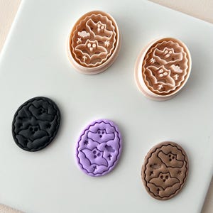 May include: Oval-shaped cookie cutters and cookies with a Halloween theme. The cutters are light brown, and the cookies are black, purple, and brown. Each features a bat and other Halloween-related shapes.