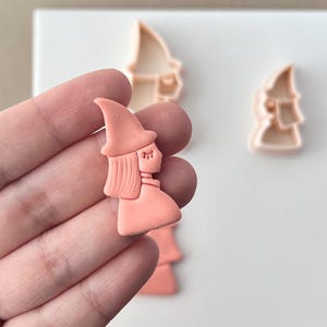 May include: A close-up of a witch-shaped cookie cutter in a light peach color. The cutter is held in a hand, with two additional cutters in the background. The witch has a pointed hat and long hair. The cutters are on a white surface.